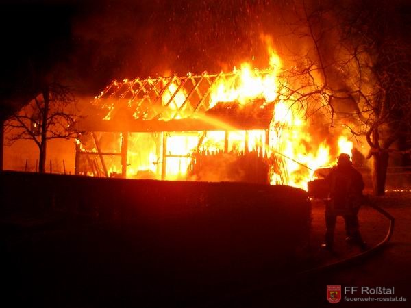 Bild 1 von 8 Die Scheune stand beim Eintreffen der Einsatzkräfte im Vollbrand. Ganz links im Bild ist ein angrenzendes Haus zu sehen, in dessen Fenster sich bereits ein kleines Feuer gebildet hat. Dies konnte rasch gelöscht werden. Rechts sieht man den PKW im Vollbrand.